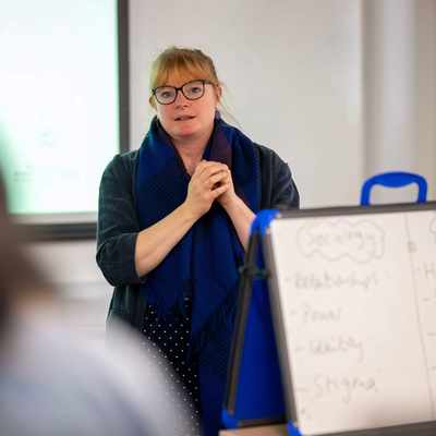 A woman wearing glasses stands in front of a whiteboard, engaged in a discussion or presentation.