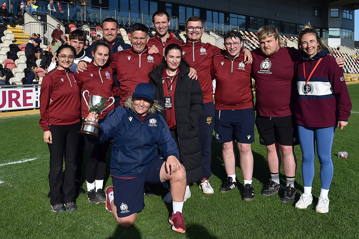 A diverse group of people posing on a rugby field, showcasing camaraderie with a bright green field behind them.