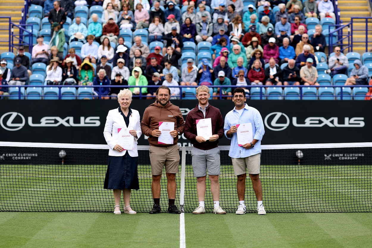 Four people stand in front of a tennis net on a court in front of a large crowd of spectators