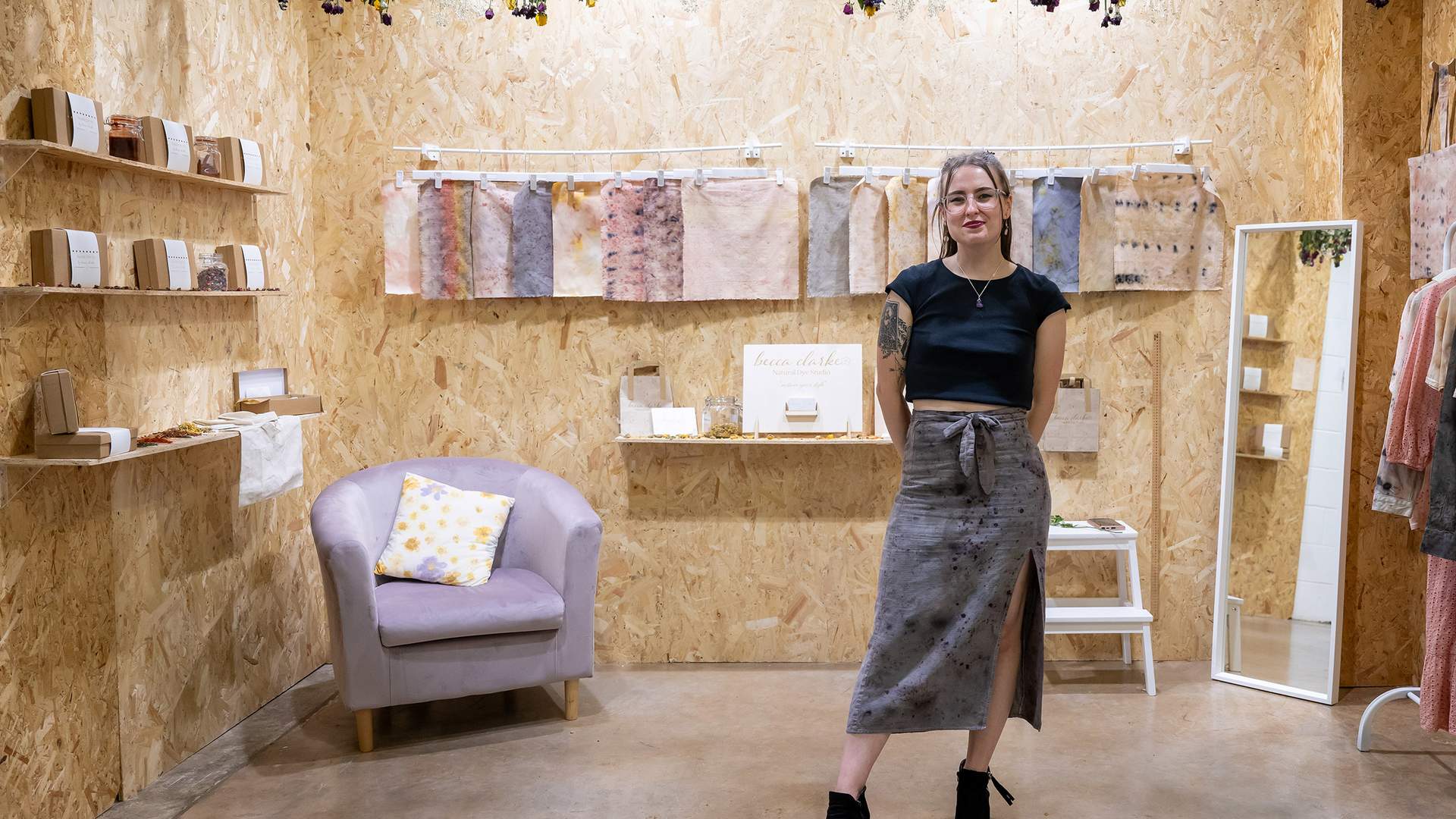 A woman stands smiling in a cozy, wood-paneled boutique. She wears a black top and a long skirt. The room features dried flowers, a chair with a patterned pillow, art on the walls, and display shelves with items.