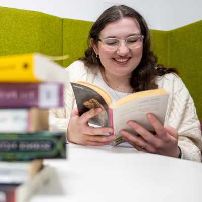 A person with glasses is smiling while reading a book. They are seated at a table with a stack of books in the foreground. The background features green upholstered panels.
