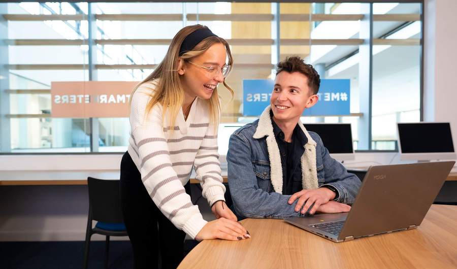 A person sits at a table in front of a laptop computer. Another person stands next to them laughing.