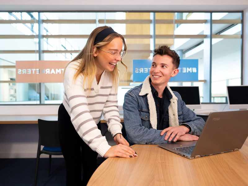 A person sits at a table in front of a laptop computer. Another person stands next to them laughing.
