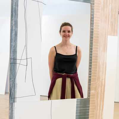 A woman stands smiling amidst an abstract art installation with geometric designs on large panels. She is wearing a black top and has a sweater tied around her waist. The setting appears to be an art gallery.
