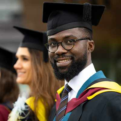 A graduate in cap and gown joyfully holds up a Cardiff Metropolitan University sign. The background is blurred, highlighting his smile and the sign.