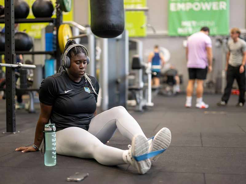 A woman sitting on the gym floor, engaged in a fitness activity, with weights and mats visible around her.