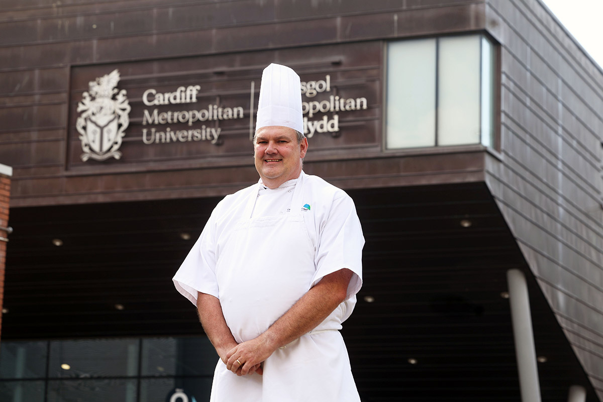 A man in a chef's uniform stands outside a building, smiling and holding a cooking utensil.