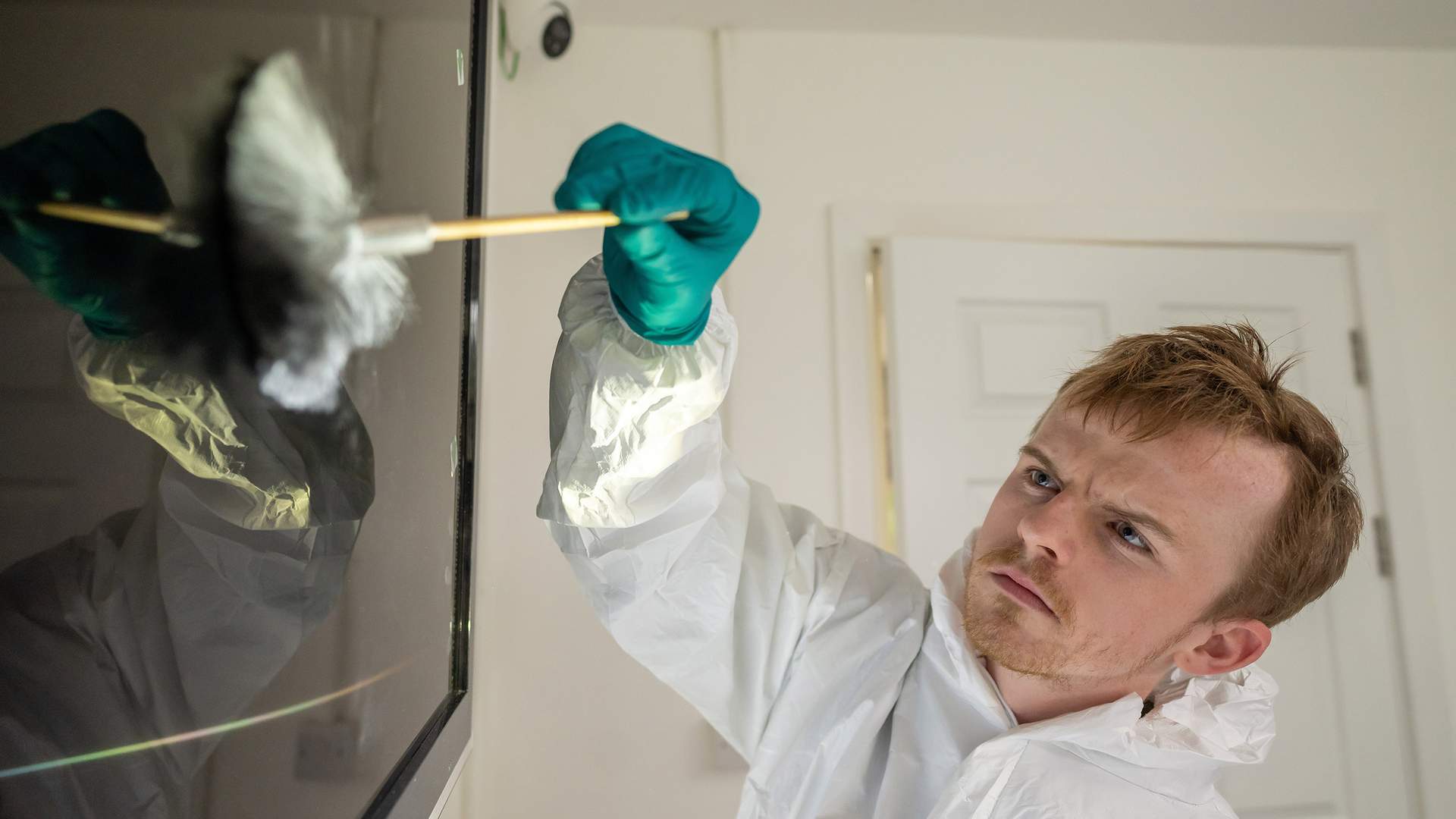 A gloved hand brushes against a reflective surface showing visible fingerprints. Green arrow markers are placed nearby. The scene is brightly lit, with lens flares visible on the left of the image.