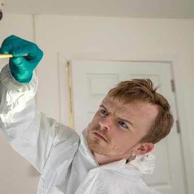 A gloved hand brushes against a reflective surface showing visible fingerprints. Green arrow markers are placed nearby. The scene is brightly lit, with lens flares visible on the left of the image.