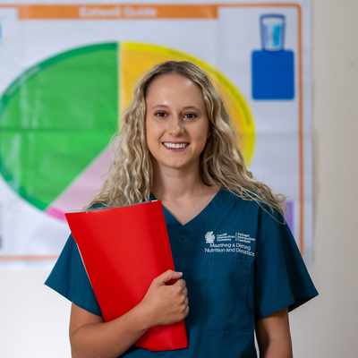 A woman in a teal uniform smiles while holding a red folder. She stands in front of a colorful chart, likely related to nutrition or dietetics. Her long blonde hair is loose, and she appears confident and professional.
