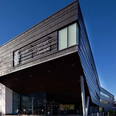 Modern building with angular architecture, featuring a large sign that reads Prifysgol Metropolitan Caerdydd. The structure has a mix of dark panels and white walls, with glass windows. Trees and a blue sky are visible in the background.