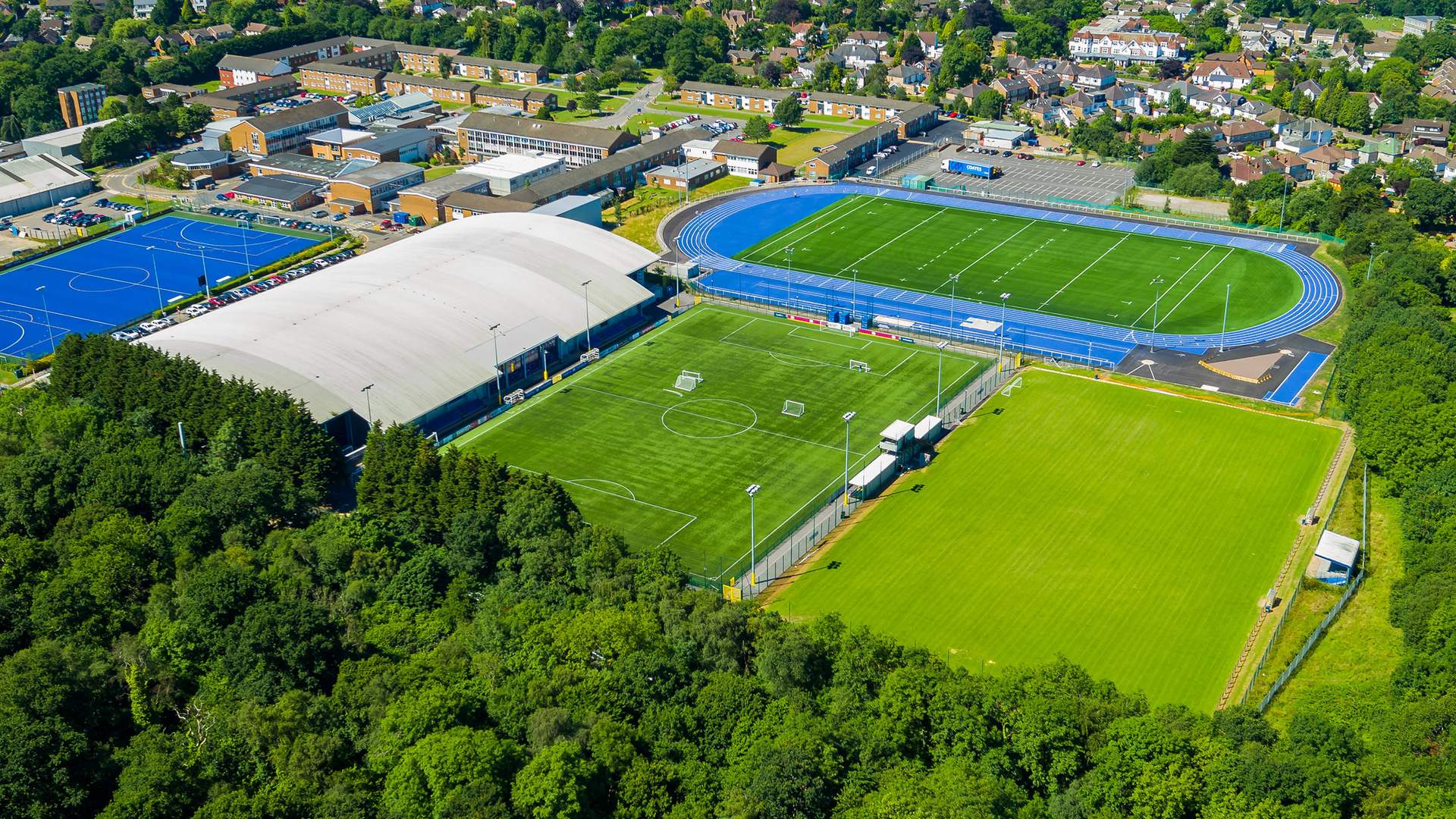 Aerial view of a sports complex featuring a large indoor sports facility, blue athletics track, multiple soccer fields, lush green trees in the foreground, and nearby residential buildings in the background.