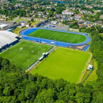 Aerial view of a sports complex featuring a large indoor sports facility, blue athletics track, multiple soccer fields, lush green trees in the foreground, and nearby residential buildings in the background.