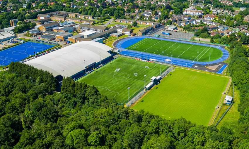 Aerial view of an athletic complex featuring a green soccer field, blue running track, and domed sports facility. Surrounding the complex are trees and residential buildings in the background.