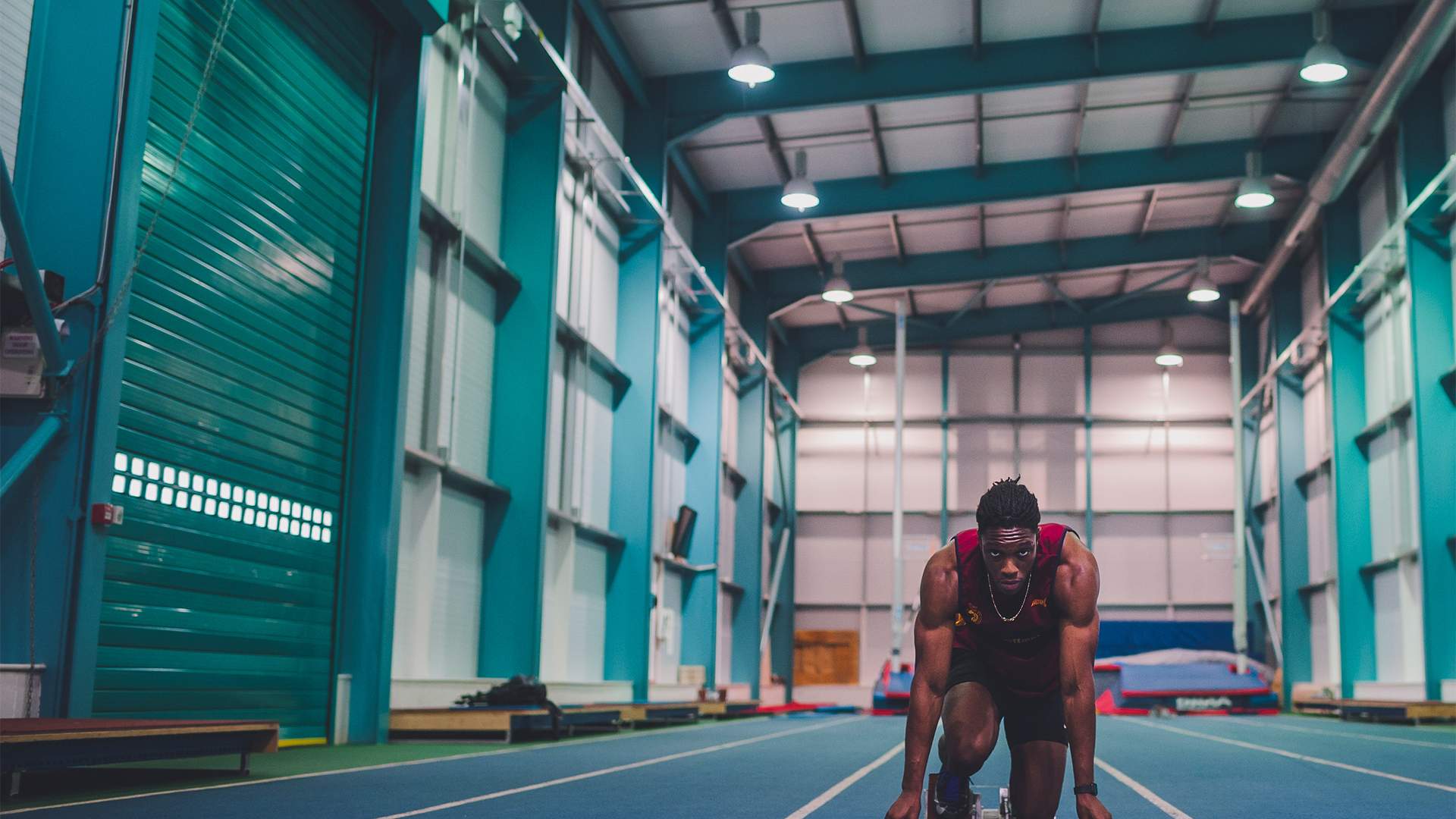 An athlete crouches in a starting position on a blue indoor track, preparing to sprint. The spacious facility is well-lit with high ceilings, teal metal beams, and closed overhead doors.