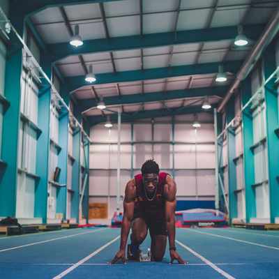 An athlete crouches in a starting position on a blue indoor track, preparing to sprint. The spacious facility is well-lit with high ceilings, teal metal beams, and closed overhead doors.