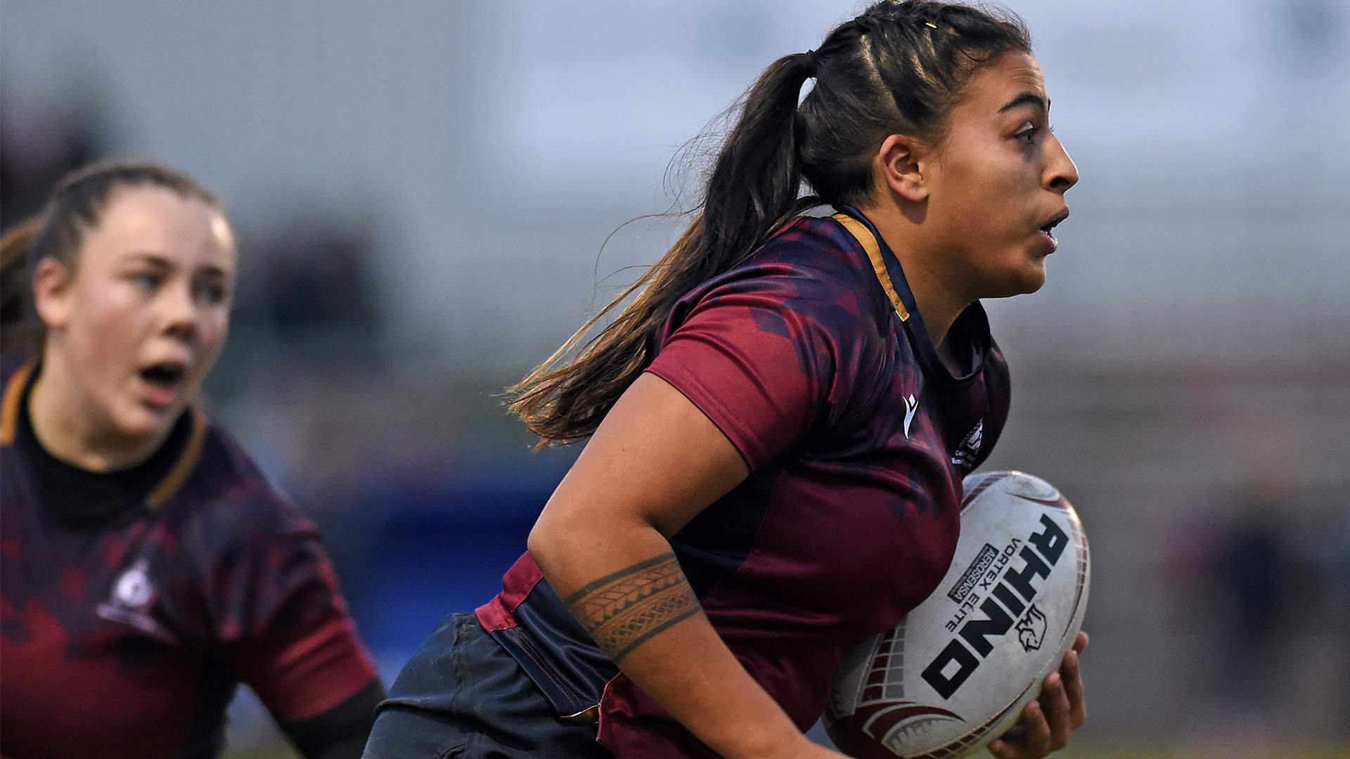 Two women in maroon jerseys engaged in a rugby match, showcasing teamwork and athleticism on the field.