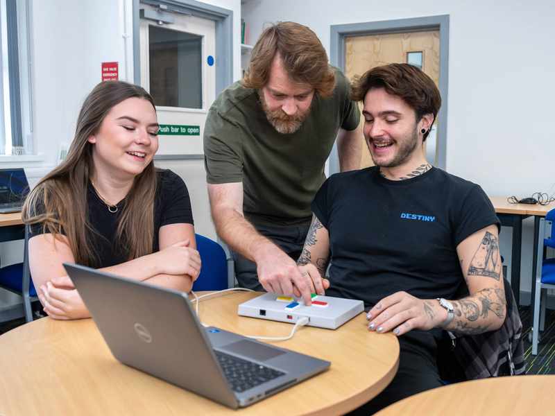 A person sits at a round table. In front of them is a small box with four buttons on it, connected to a laptop computer.