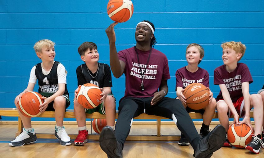 A sports coach spins a basketball on his fingers while four pupils, all holding a basketball on their laps, watch on