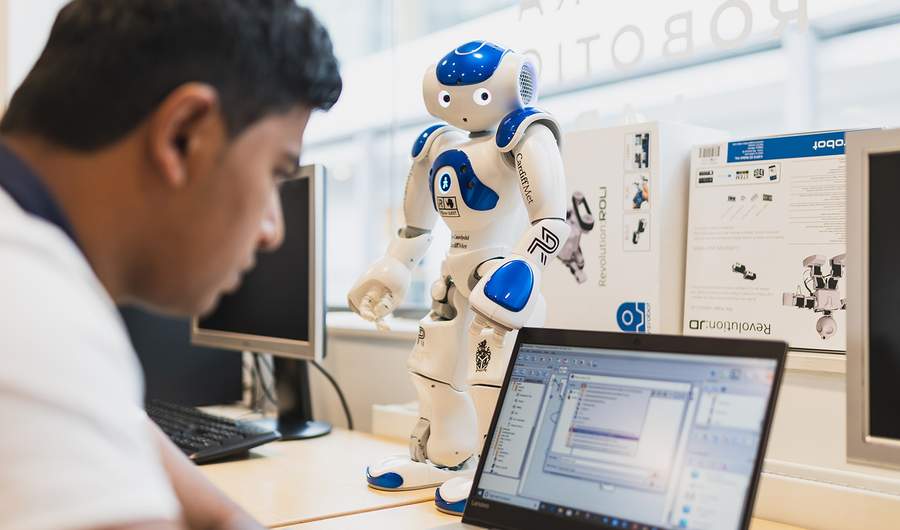 A man focuses on his laptop while observing a small robot beside him, engaged in a technological task.
