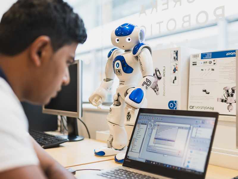 A man focuses on his laptop while observing a small robot beside him, engaged in a technological task.