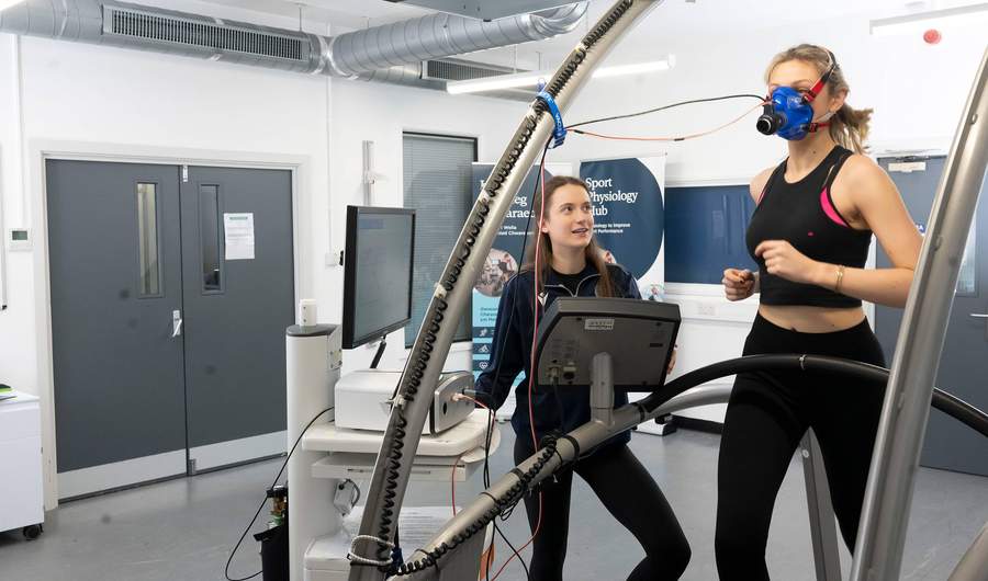 A person runs on a treadmill while wearing a breathing mask. A second person stands alongside, next to a computer display and other equipment.
