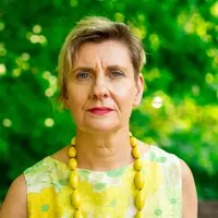 A person wearing a flower pattern top and a necklace with large yellow beads. In the background is some green foliage.