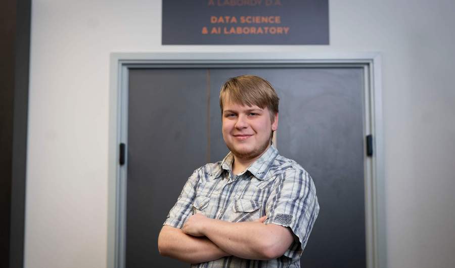 A person stands with arms folded in front of a door. Above the door is a sign that reads Data Science and AI Laboratory.
