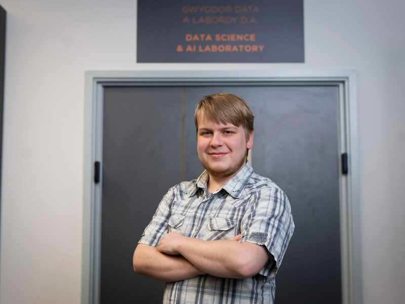 A person stands with arms folded in front of a door. Above the door is a sign that reads Data Science and AI Laboratory.