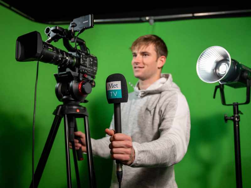 A person stands behind a television camera. Behind them is a green backdrop.