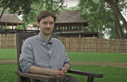 A young man sits on a wooden chair in an open green area