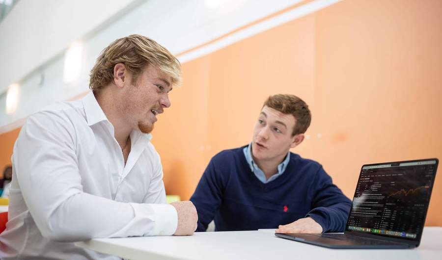 Two people sit next to each other. On the table in front of them is a laptop computer.