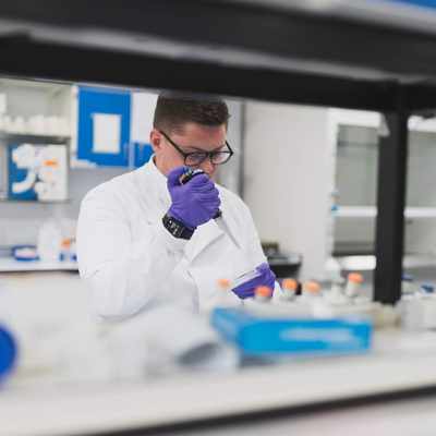 A person in a lab coat uses a pipette to dispense a sample into a container.