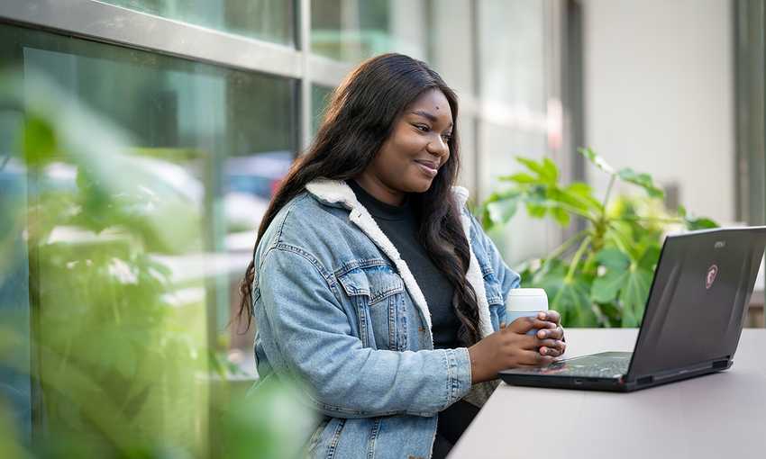A young woman sits at an outdoor table holding a coffee in both hands while reading from their laptop screen