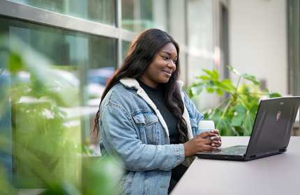 A young woman sits at an outdoor table holding a coffee in both hands while reading from their laptop screen
