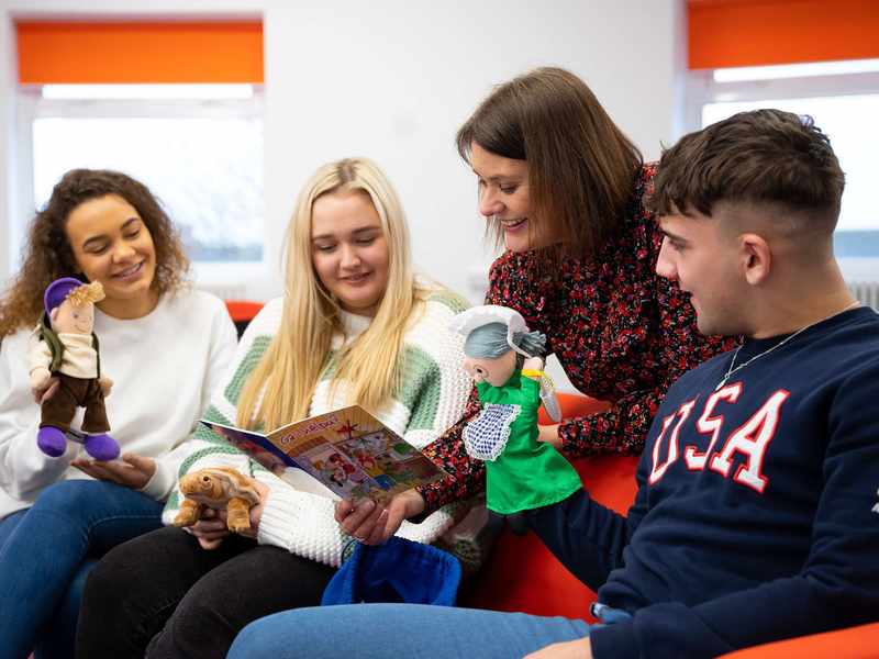 A person reads a children's book while three other people holding puppets and toys look on.