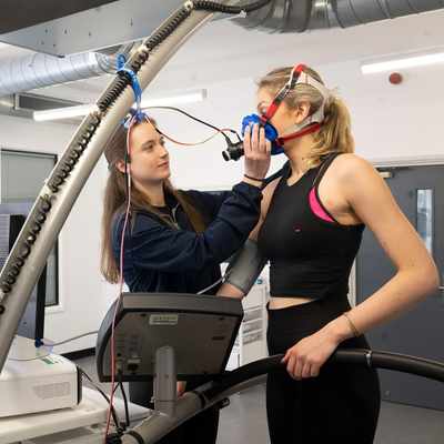 A person attaches a breathing mask to another person, who is standing on a treadmill. In front of them are computer monitors.