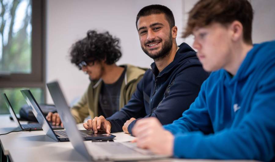 Three people sit alongside each other at a row of desks. Each has a laptop computer in front of them.