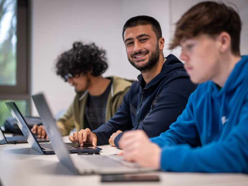 Three people sit alongside each other at a row of desks. Each has a laptop computer in front of them.