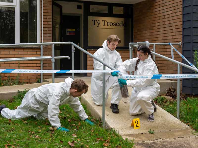 Three students wearing white jumpsuits and gloves inspect a fake crime scene for clues