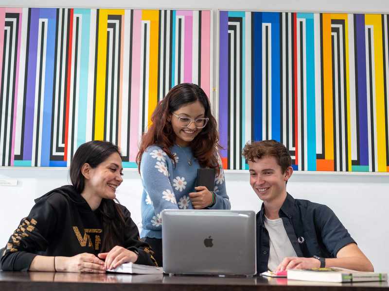 Two people sit at a table in front of a laptop. Behind them stands a third person.