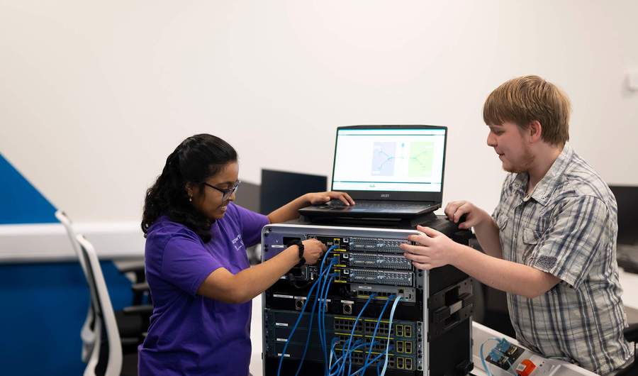 Two people plug cables into computer network equipment.