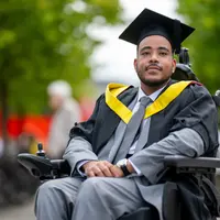 A man in a graduation gown and cap sits in a wheelchair, smiling proudly at his achievement.