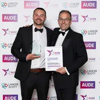Two men in suits proudly hold an award in front of a decorative wall.