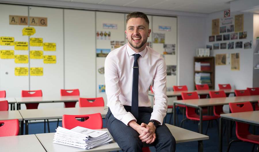 A teacher sits on a desk in an empty classroom.