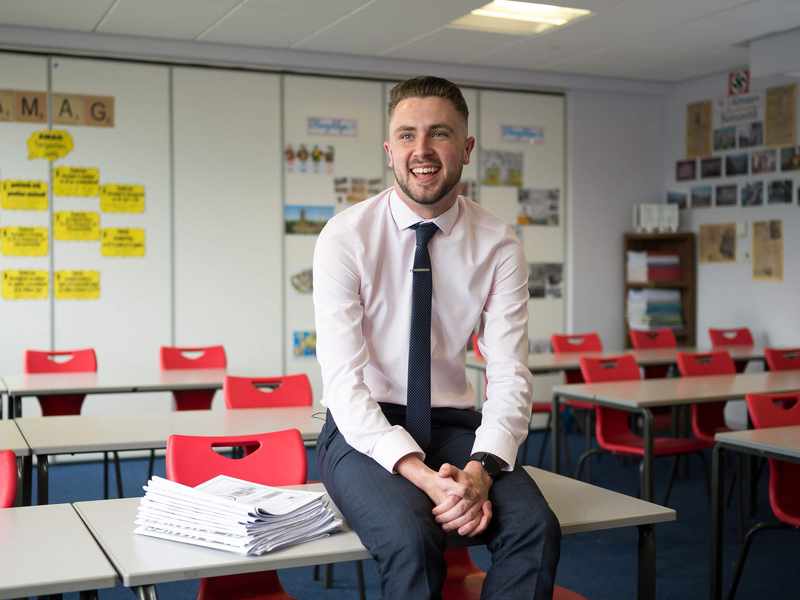 A teacher sits on a desk in an empty classroom.