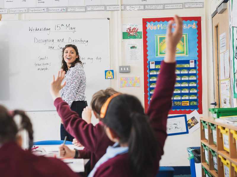 A teacher writes on a whiteboard while two pupils raise their hands.