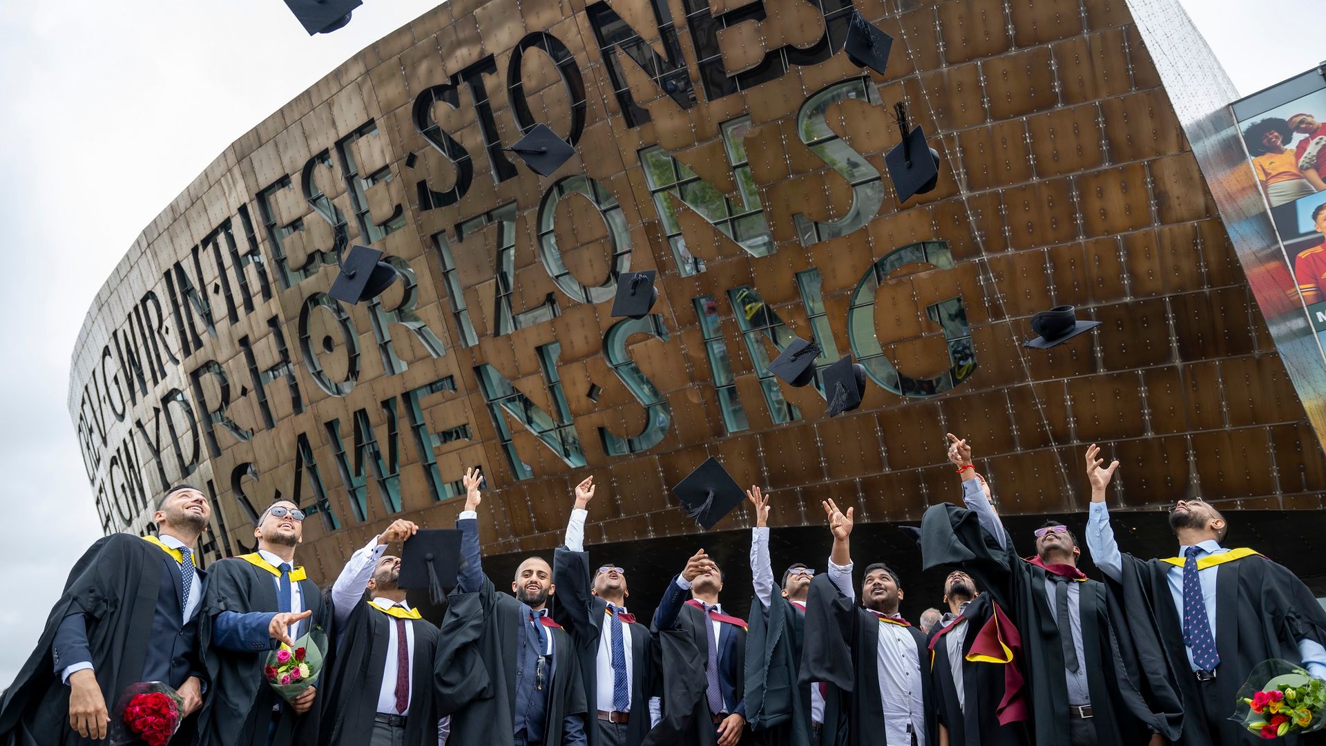 A row of student toss their graduation caps into the air in front of the Wales Millennium Centre sign and entrance