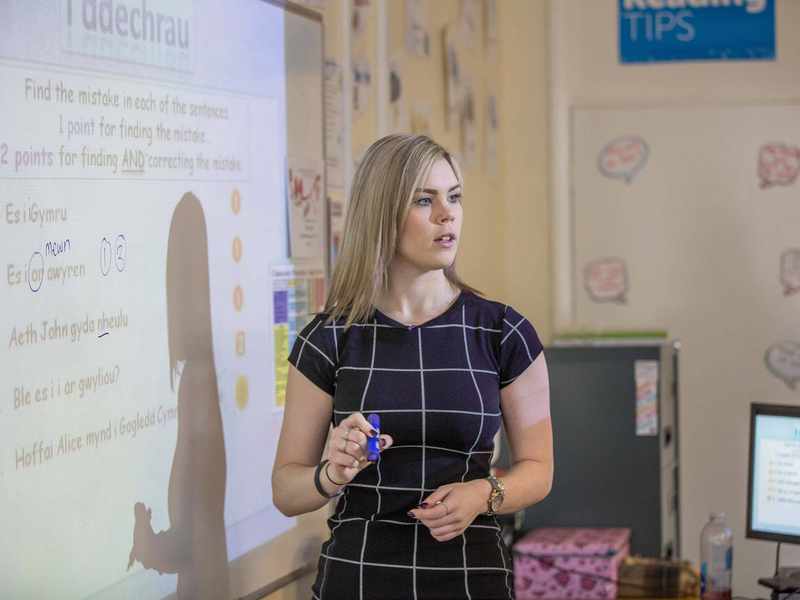 A teacher stands next to a whiteboard. Written on the whiteboard is some text in Welsh.
