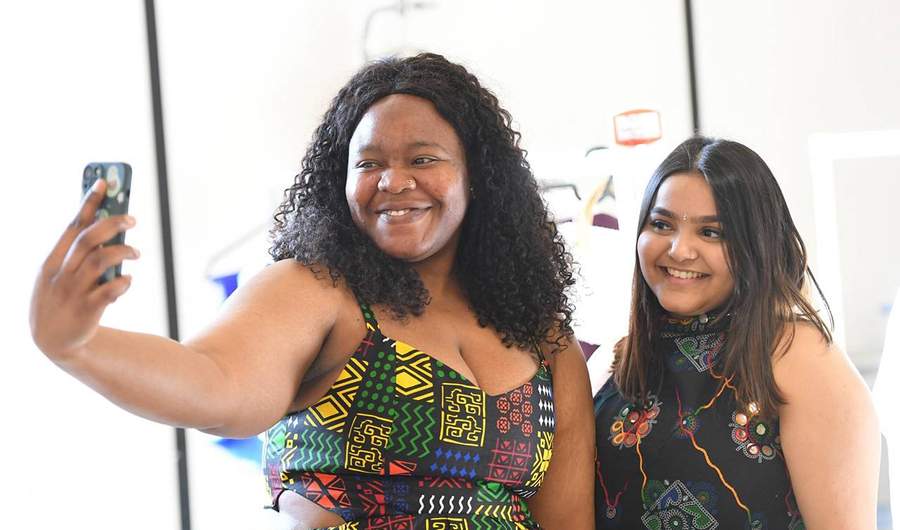Two people pose for a selfie. They are wearing black dresses, patterned with bright colours.